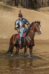 A young adult man in knightly armor rides a horse on a river along a sandy shore