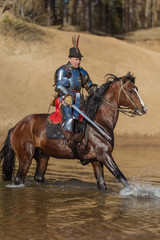 A young adult man in knightly armor rides a horse on a river along a sandy shore