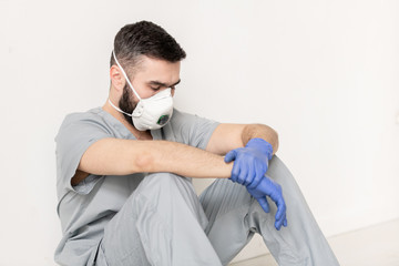Tired doctor in gloves, respirator and grey uniform sitting on the floor by wall