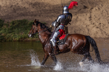 A young adult man in knightly armor rides a horse on a river along a sandy shore