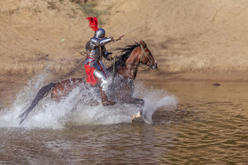 A young adult man in knightly armor rides a horse on a river along a sandy shore