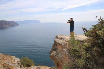 A man, a tourist stands on a high mountain and photographs the landscape, the sea on the phone. Beautiful sea coast, view from the cliff. Traveling alone. The concept of beautiful views of nature.