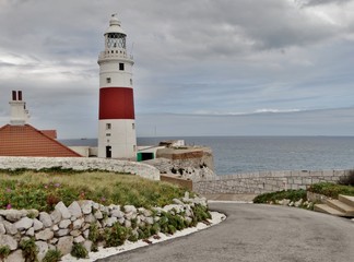 Fotografía de un faro de color blanco y franja de color rojo, antiguo,  al final de un camino y con el Mar Mediterráneo al fondo en el peñón de Gibraltar