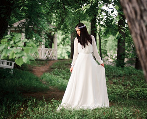 A beautiful bride in a summer green garden against the background of a vintage white gazebo