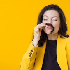 Stylish young woman in a yellow jacket on a yellow background, making a mustache out of her hair.