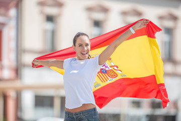 Happy smiling pretty girl holds a flag of Spain behind her celebrating on a street