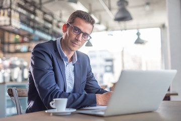 Young entrepreneur wearing glasses looks at the camera during a coffee break in a cafe with a computer in front of him