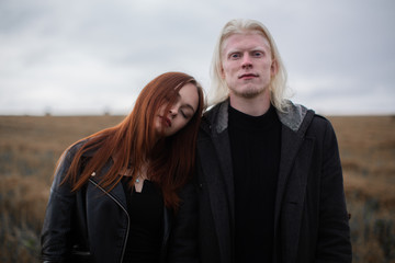 A red-haired girl is lying on the shoulder of an albino guy. The pair are standing in the field. The guy looks into the camera, the girl with his eyes closed