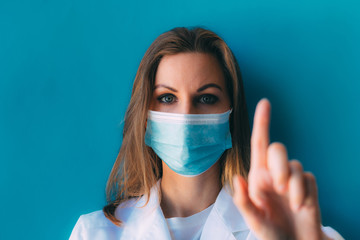 Portrait of young female doctor in medical mask and white gown on blue background. Female doctor in clinic in surgical mask with space for text. Looking on camera and shows hand with finger up