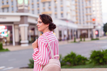 Fototapeta premium Portrait of a cute, beautiful girl 30-33 years old in the city. Young woman in a beautiful pink dress with makeup and hairstyle. On the stove of a woman is a cute backpack.