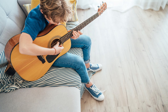 Preteen boy playing acoustic guitar dressed casual jeans, shirt and new sneakers sitting on the cozy sofa at home living room. Music education top angle view concept image.
