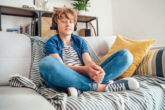 Preteen boy sitting cross-legged at home on cozy sofa dressed casual jeans and new sneakers listening to music using wireless headphones and smiling looking in camera