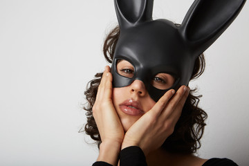Close-up of young woman with bunny ears mask, raises palm holding her head, gestures over blank...