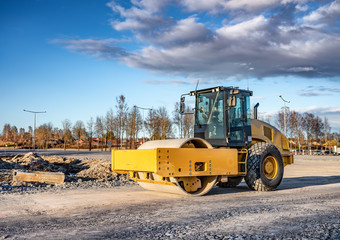 Eye catching yellow road roller with enclosed climate controlled cabin stands on not ready new road, stones, blue sky, clouds, front right side view. Clean shiny old heavy tractor