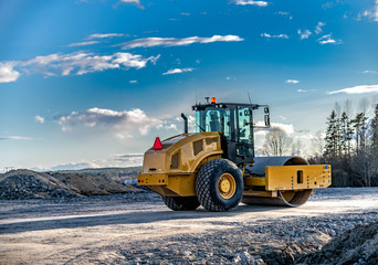 Eye catching yellow road roller with enclosed climate controlled cabin stands on not ready new road, stones, blue sky, clouds, back left side view. Clean shiny old heavy tractor