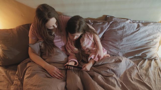 Mother daughter bonding. Family leisure. Woman girl watching movie on phone together in bed.