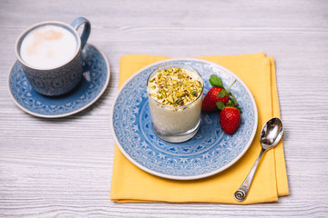Pistachio parfait in a transparent glass cup on a blue plate with two strawberries, on a yellow napkin with a teaspoon. A cup of cappuccino stands on the side.