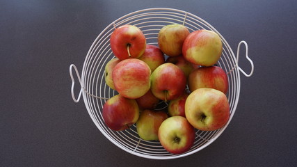 primer plano de  un frutero blanco con asas en forma de corazon  lleno de manzanas rojas con un fondo gris