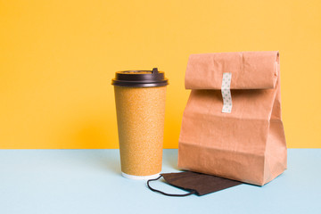 paper cup of coffee and bag for delivery, yellow with blue background, copy space, black protective mask for courier, face mask as a gift when ordering