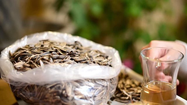 A woman eats dry and salty Sunflower Seeds and drinks green tea in her balcony or backyard. Green plants and small fountain at background.