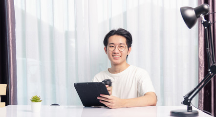 Happy Asian young business handsome man work from home office wear glasses, t-shirt comfortable he smiling and using a black modern smart digital tablet computer to read email or learning on desk
