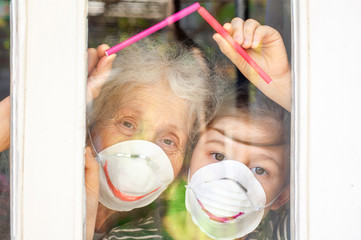 Grandmother with grandchild in a respiratory masks plays with the window of his house. Family fun....