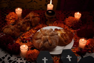 Pan de Muerto, traditional and sugared, around it marigold flowers, cardboard coffin, lighted candles, lit copal, diffused confetti background.