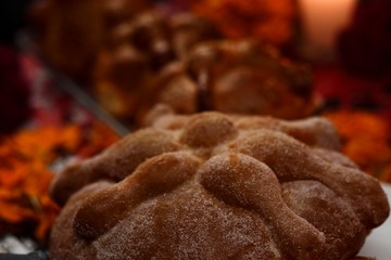 Pan de Muerto sugared, placed on a glass plate, around marigold flowers, burning candles, in the background traditional pan de Muerto