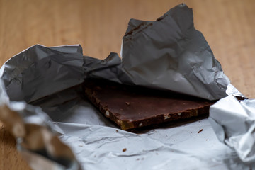 An unwrapped chocolate bar with a corner block of dark chocolate with nuts. The silver wrapper and package is torn open and laying on a wooden table. The sweet dessert snack has antioxidants. 