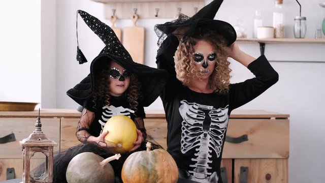 Woman and girl wear costumes. Mother and daughter have black hats. Girls play together in kitchen.