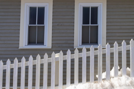 White Wooden Picket Fence In The Foreground With A Tan Colored Wooden Clapboard Exterior Of A House. The Building Has Two Double Hung Vinyl Windows In The House. There's A Drift Of Snow By The Fence. 