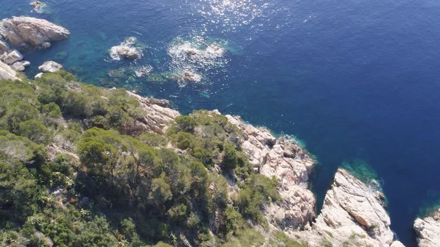 Aerial Drone side close-up view of mediterranean cliff with green trees, rocks and blue water