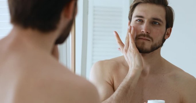 Attractive shirtless metrosexual young guy applying moisturiser cream on face looking in mirror. Handsome bearded man holding creme jar doing morning skin care beauty routine. Male skincare concept
