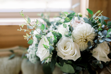 A Beautiful Arrangement of Flowers with White Roses, Carnations, and Eucalyptus Leaves and Green Olive Leaves near an open window.  This is a wedding bouquet. 