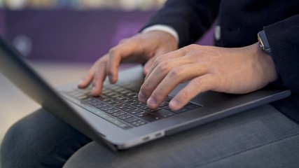 Man s hands with smartwatch typing on grey keyboard of modern laptop lying on knees. Close up of young businessman hands dressed in suit typing on keyboard