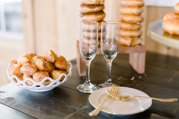 Two champagne flutes sit on a wood table with a bowl of glazed donut holes and a plate with two gold forks. A stack of donuts are in the background. 