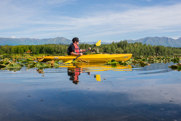 Teenager paddles a bright yellow canoe in a tranquil lily pad filled Alaskan lake.