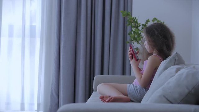 Curious cute girl preschool child using a digital tablet technology device looks telephone.Plays in the phone sitting on the sofa alone.Children tech addiction concept play game at home.