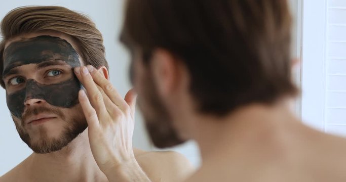 Smiling young bearded metrosexual man applying charcoal clay facial mask looking in bathroom mirror. Handsome shirtless guy cleaning face skin with spa cleansing mud. Male skincare beauty treatment.