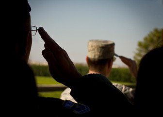 Close up of man with glasses in silhouette saluting with another man in background in camouflage hat in sunlight saluting