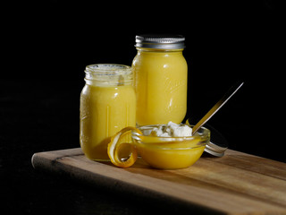 Homemade lemon curd served in a glass bowl with whipped cream and a lemon peal slice on wood cutting board agains a black background.