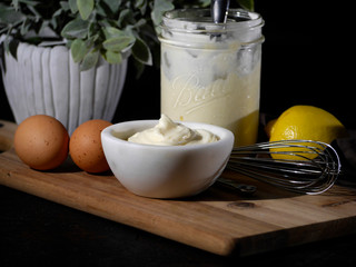 White bowl of homemade mayonnaise displayed with ingredients including brown eggs and a fresh lemon in a jar on a wood cutting board against a black background. 