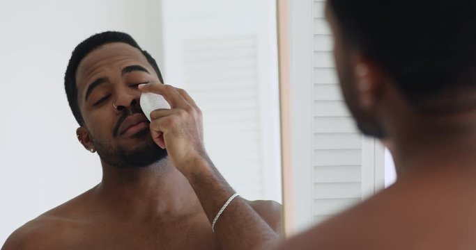 Handsome young shirtless african cleaning purifying facial skin care holding cleansing exfoliating sponge looking in bathroom mirror. Attractive ethnic guy doing skincare peeling treatment concept.