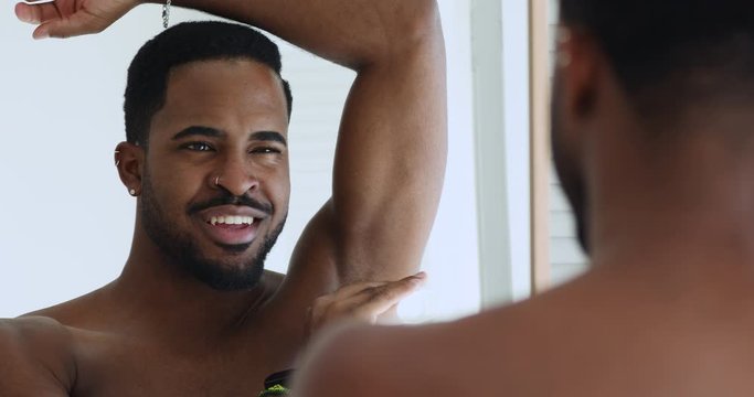 Smiling sexy shirtless african american young man applying deodorant antiperspirant stick in armpit looking in bathroom mirror. Male daily hygiene hyperhidrosis treatment sweat protection concept.