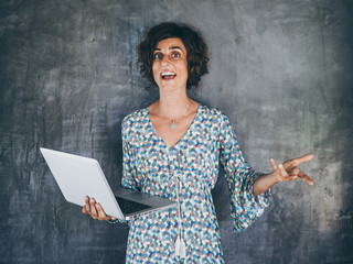 happy smiling remote digital nomad woman in colorful casual dress standing in front of a grey...