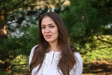 Portrait of a young brown-haired beautiful girl with green eyes and a white blouse on a background of trees