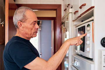 Old man with grey hair using the microwave oven in his kitchen