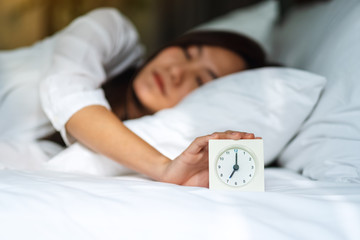 An asian woman turning off an alarm clock while sleeping on a white cozy bed in the morning