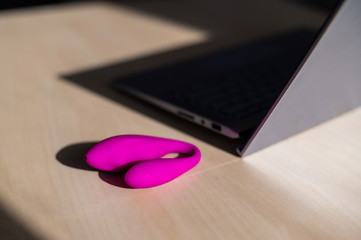Dildo on the table next to a laptop. The concept of online sex. Curved pink vibrator for female self-satisfaction. Stimulator for the clitoris.