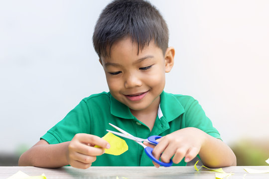 Portrait image of 5-6 years old Asian child boy practice to cutting color paper by the scissors on the wooden table.Study​ from​ home, social​ Distancing, Kid and education concept.